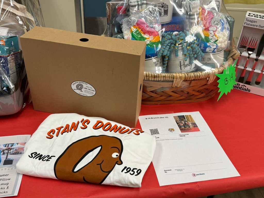 A display featuring a brown box labeled 'Stan's Donuts' next to a white t-shirt with the same branding, marked 'Since 1959'. In the background, there is a basket filled with various items, including kitchen utensils and a sign indicating 'B-14'. A sheet of paper details an auction item titled '8-14 Brunch (rm 12)'.